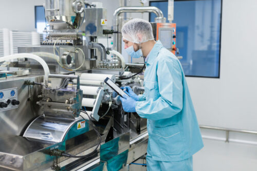 caucasian scientist in blue lab uniform configure manufacture machine with shafts and control panel, checking readings, making notes